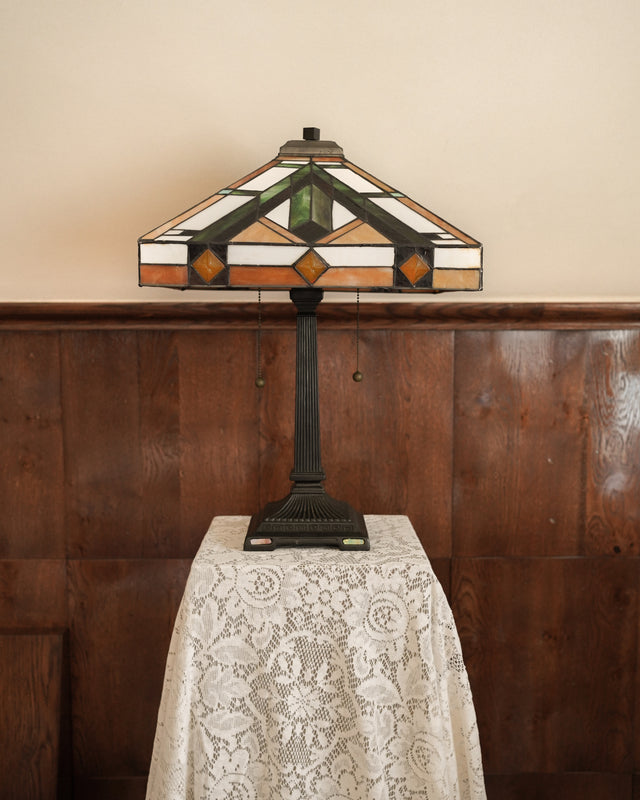 Decorative table lamp with stained glass shade on a lace-covered table against a wooden panel wall.